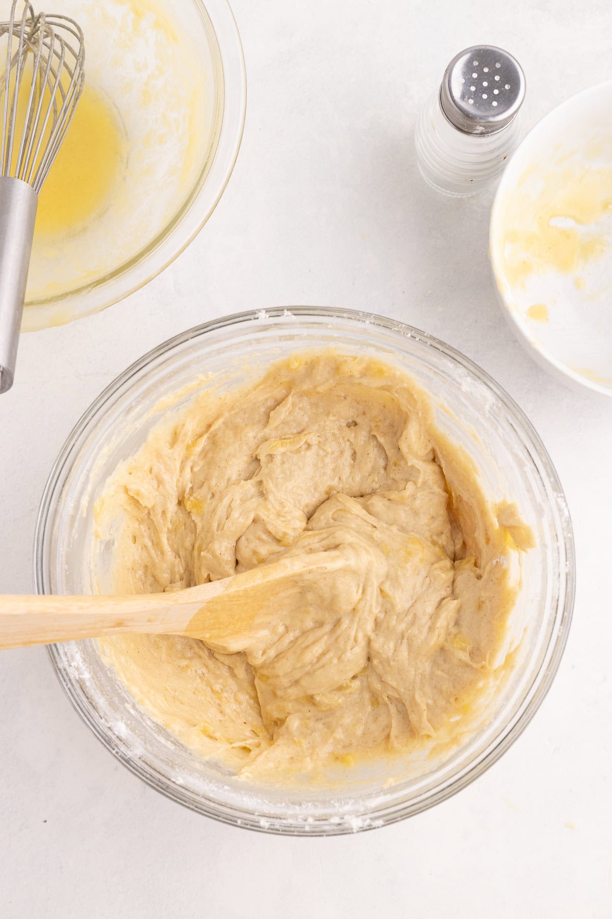 A glass bowl with thick batter being mixed with a wooden spoon, surrounded by a whisk, a salt shaker, and a small bowl on a white surface.