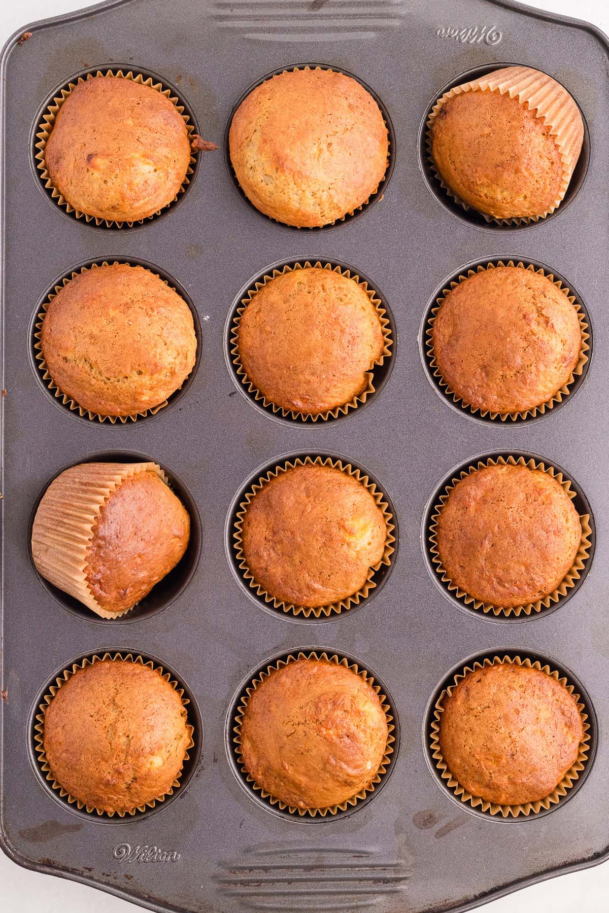 A muffin tin holds twelve baked banana muffins, some with slightly tilted paper liners, viewed from above.