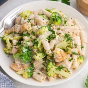 A plate of penne pasta with broccoli, grilled chicken pieces, and creamy white sauce, garnished with chopped herbs.