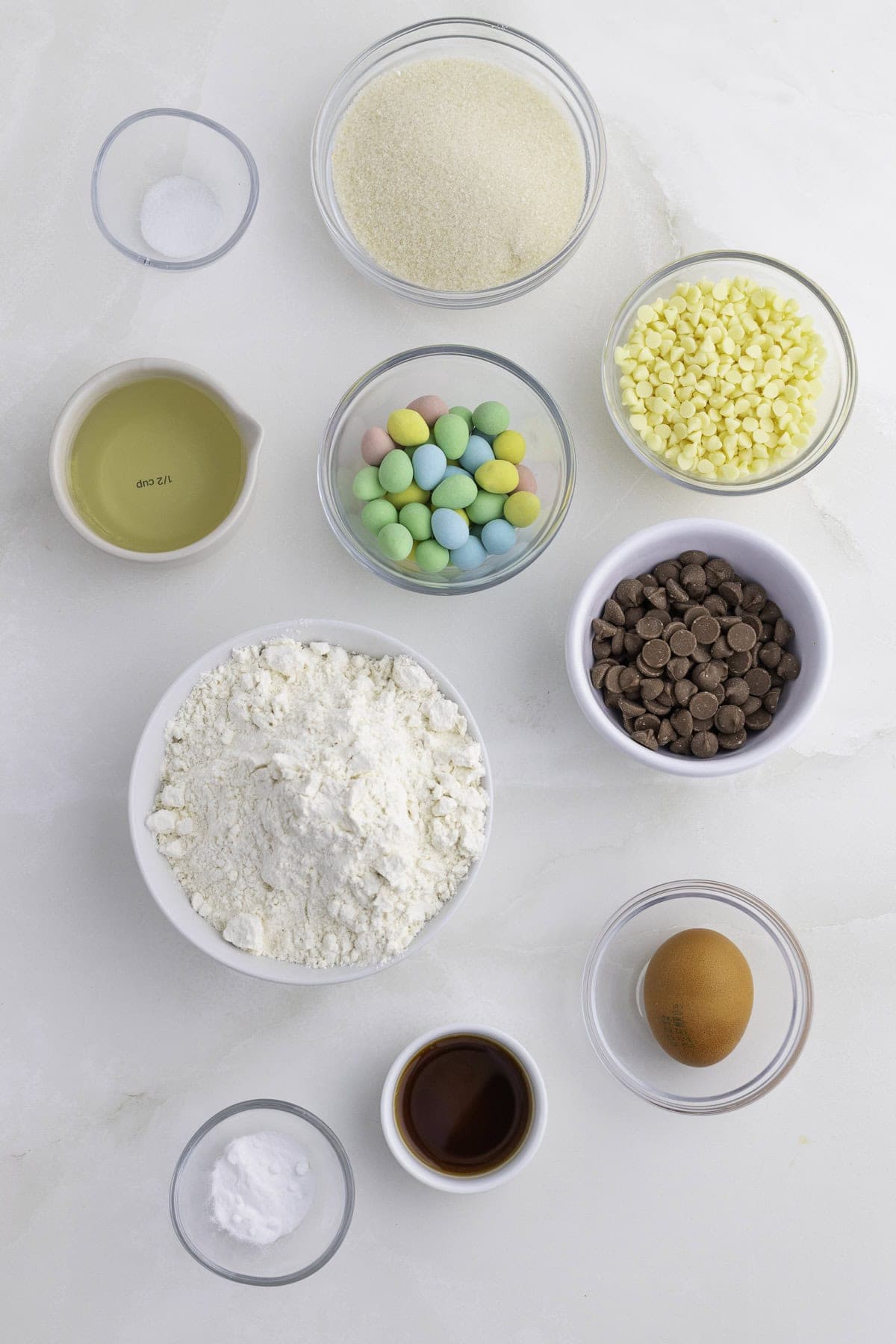 A flat lay of baking ingredients including flour, sugar, salt, egg, vanilla, oil, baking soda, white and milk chocolate chips, and pastel candy-coated chocolates in glass bowls.