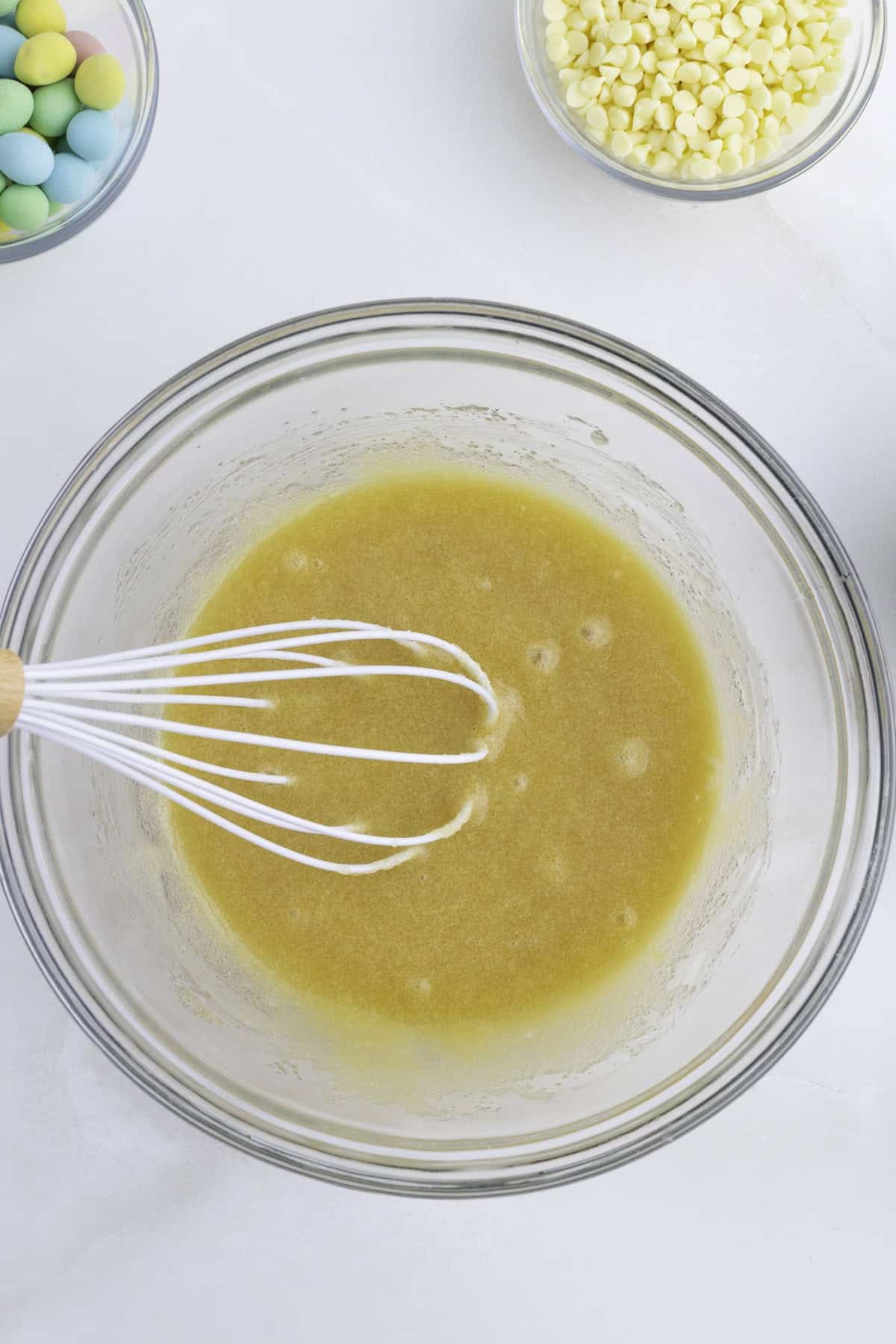 A hand holding a whisk mixes a yellow batter in a glass bowl; bowls of white chocolate chips and colorful candy are nearby on a white surface.
