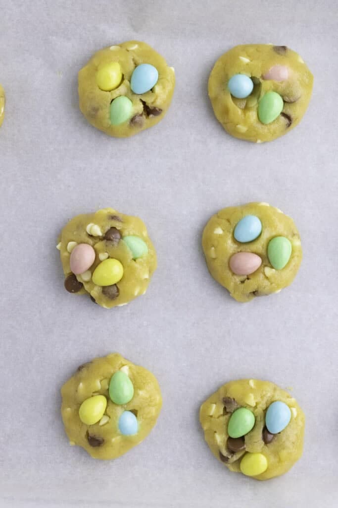 Rows of unbaked cookies with chocolate chips and colorful candy-coated eggs arranged on a sheet of parchment paper.