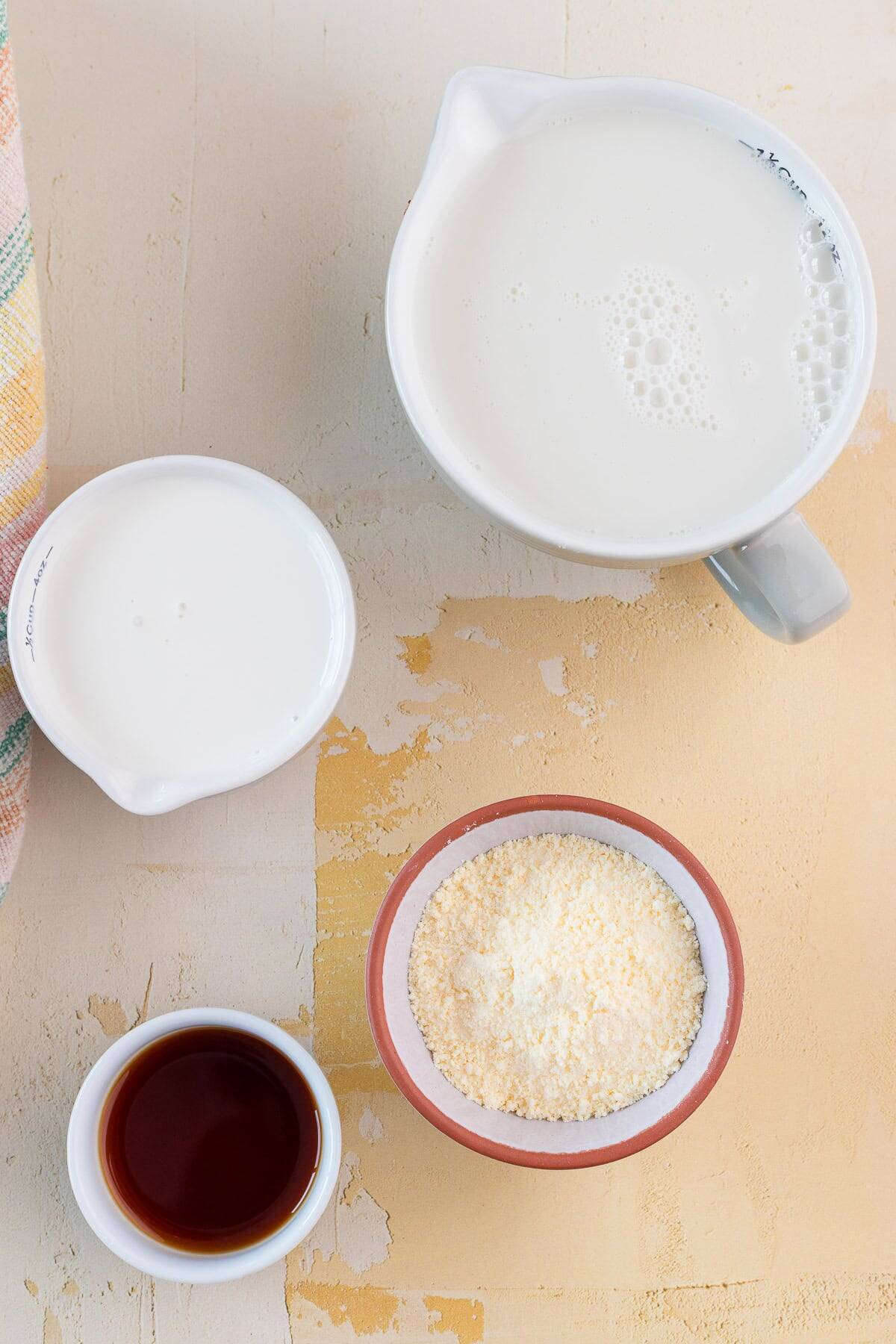 Four containers on a light surface: one with milk, one with cream, one with protein powder, and one with vanilla extract.
