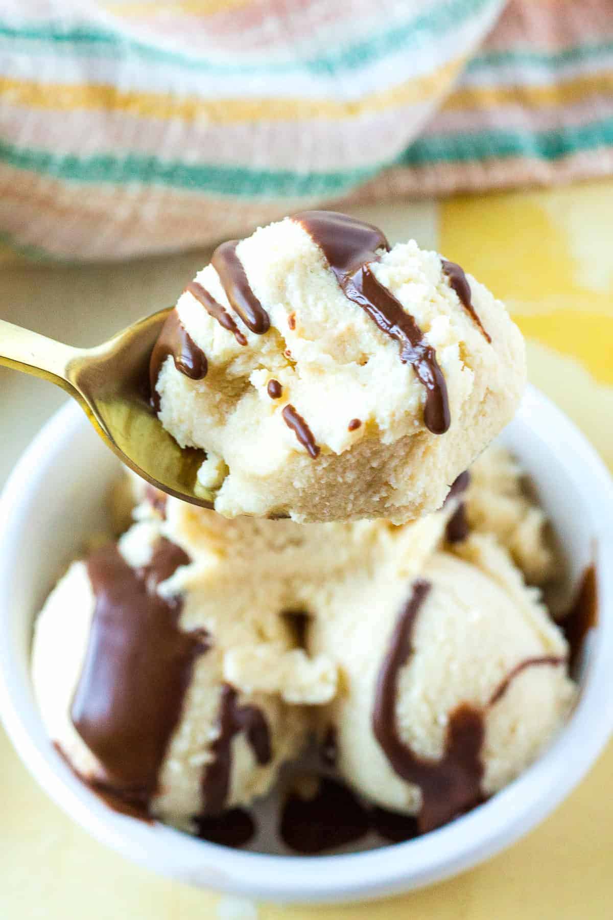 A spoonful of vanilla ice cream with chocolate syrup held above a bowl containing more scoops, topped with chocolate syrup.