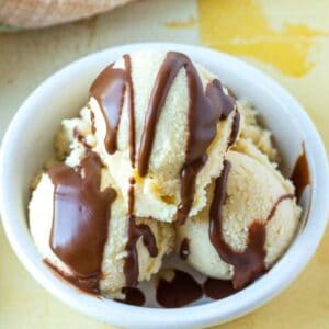 Three scoops of vanilla ice cream in a white bowl topped with chocolate sauce.