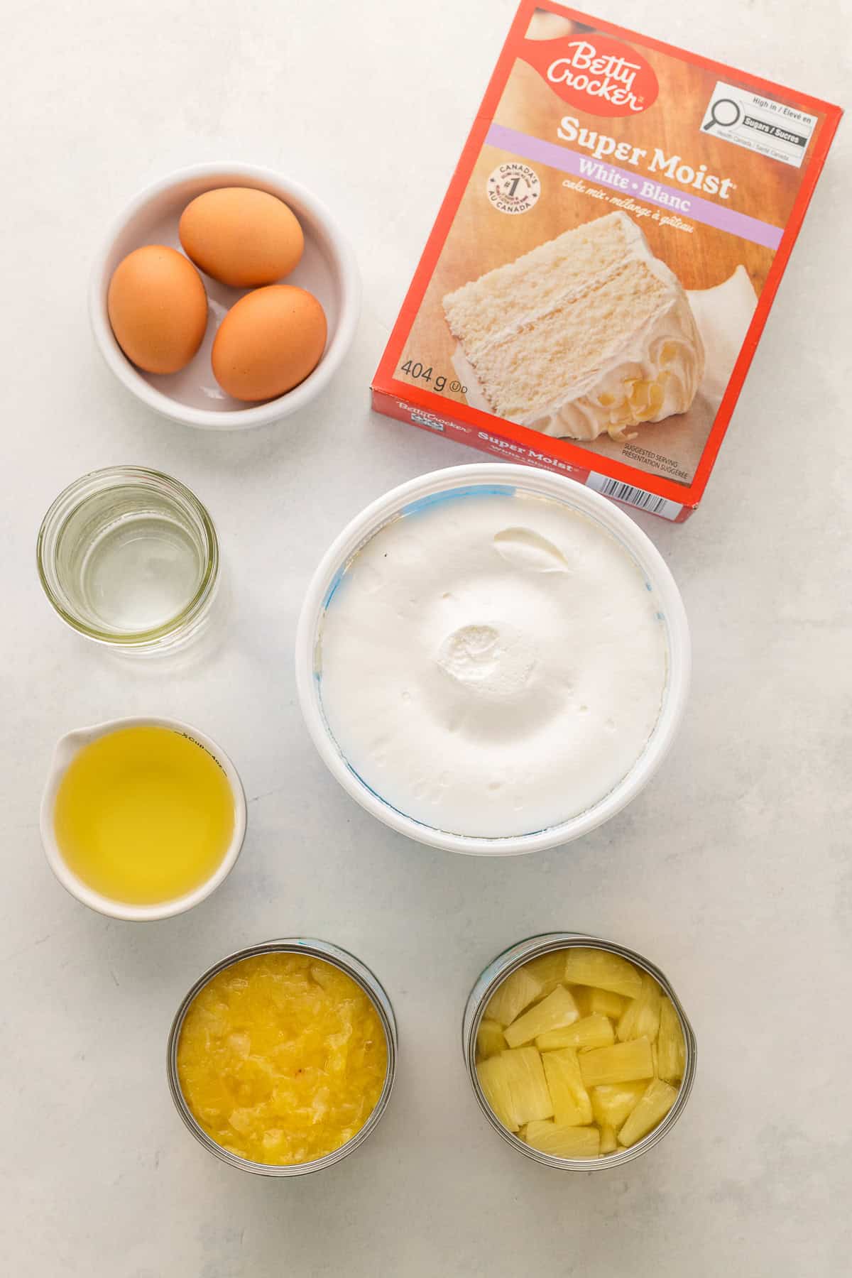 Ingredients for a cake recipe: three eggs, boxed white cake mix, vegetable oil, whipped topping, water, crushed pineapple, and pineapple chunks on a white surface.