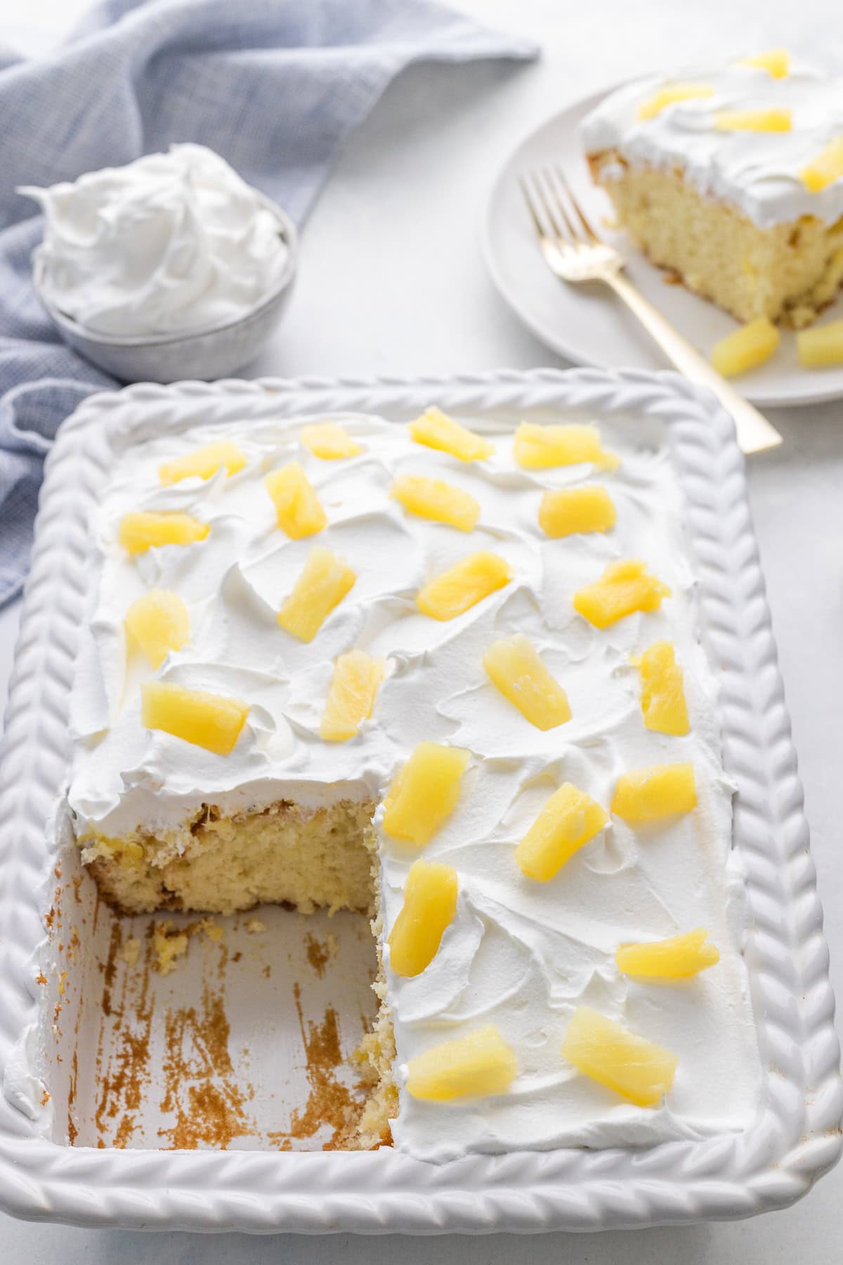 A rectangular cake topped with whipped cream and pineapple chunks in a white dish, with one slice served on a plate in the background.