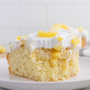 A slice of pineapple poke cake topped with white whipped cream and pieces of pineapple, served on a white plate.