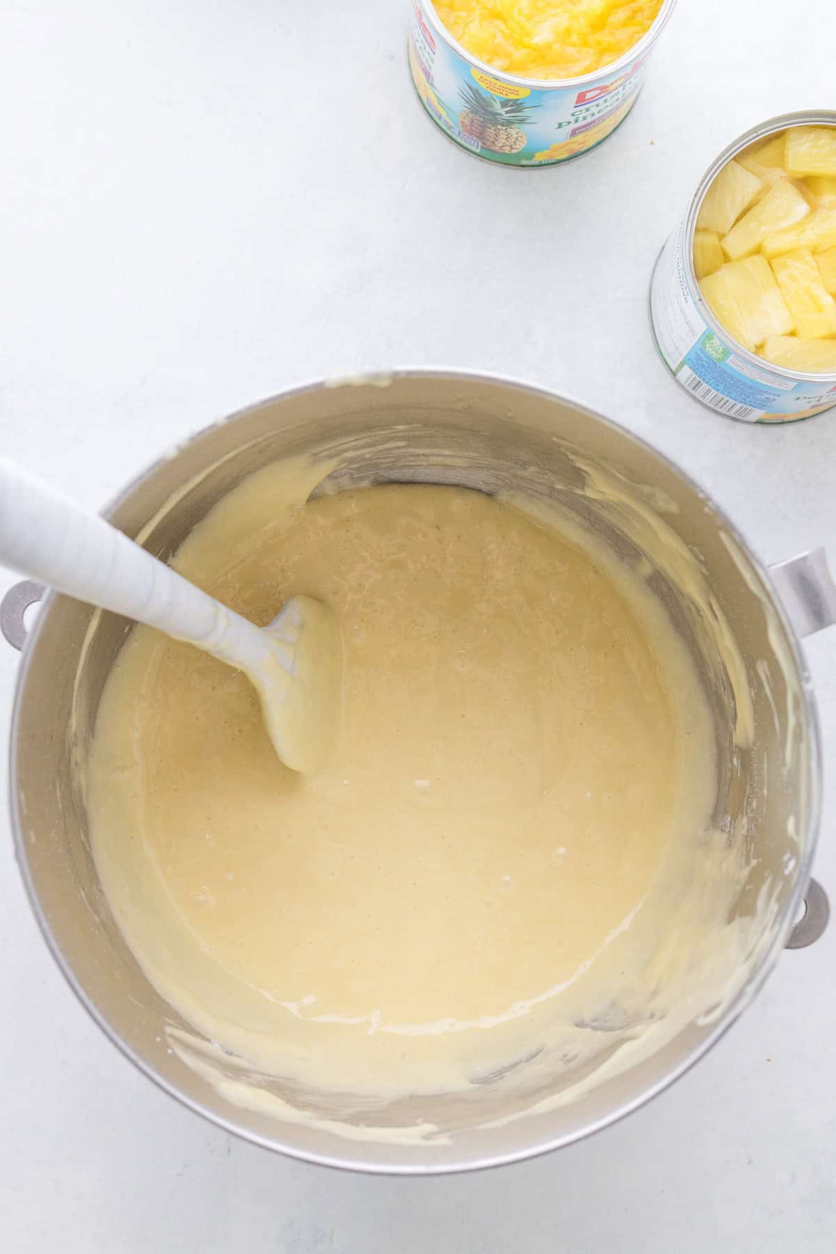 A mixing bowl with cake batter and a spatula, next to two open cans of pineapple on a light countertop.