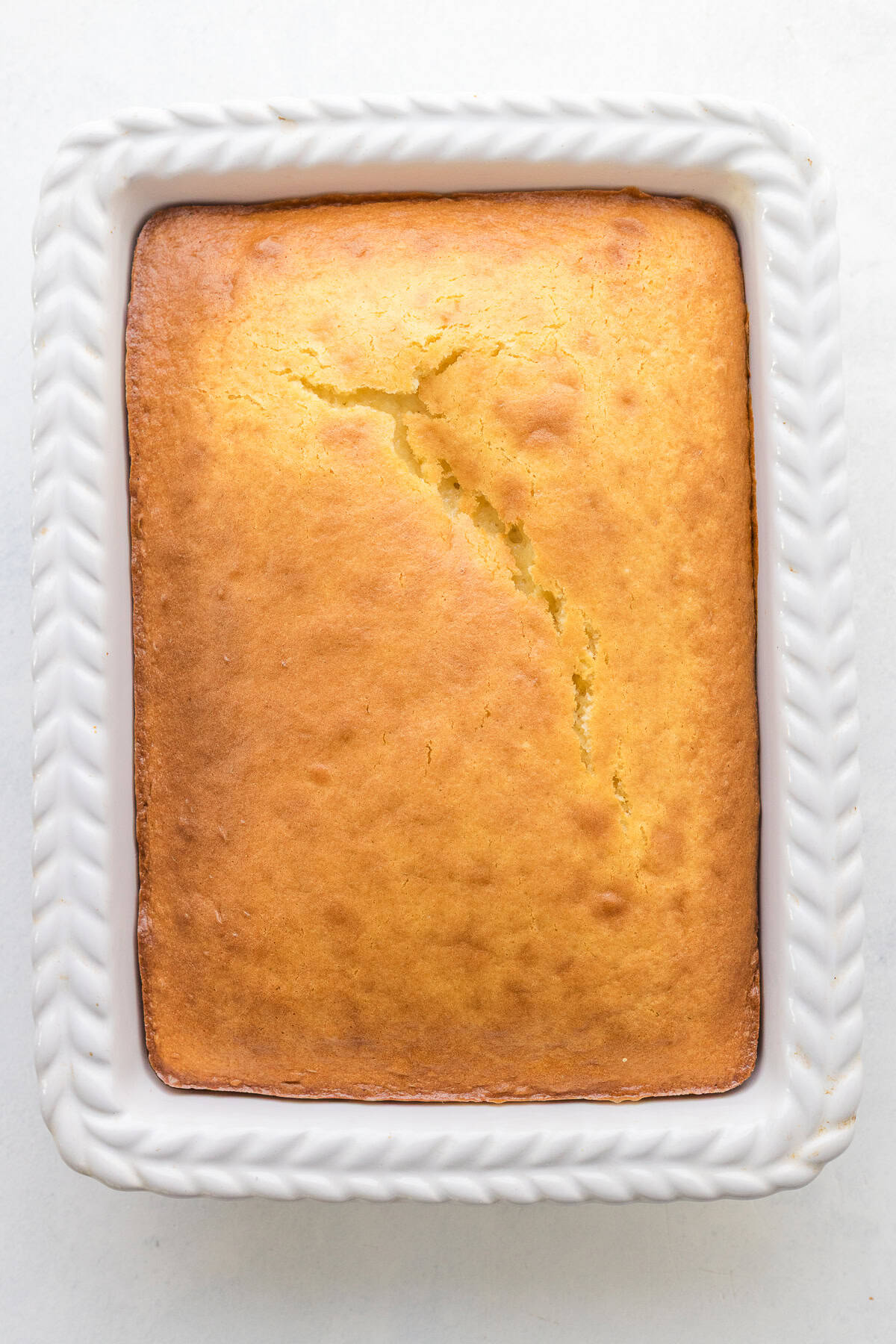 A golden-brown rectangular vanilla cake with a crack in the center sits in a white, patterned ceramic baking dish.