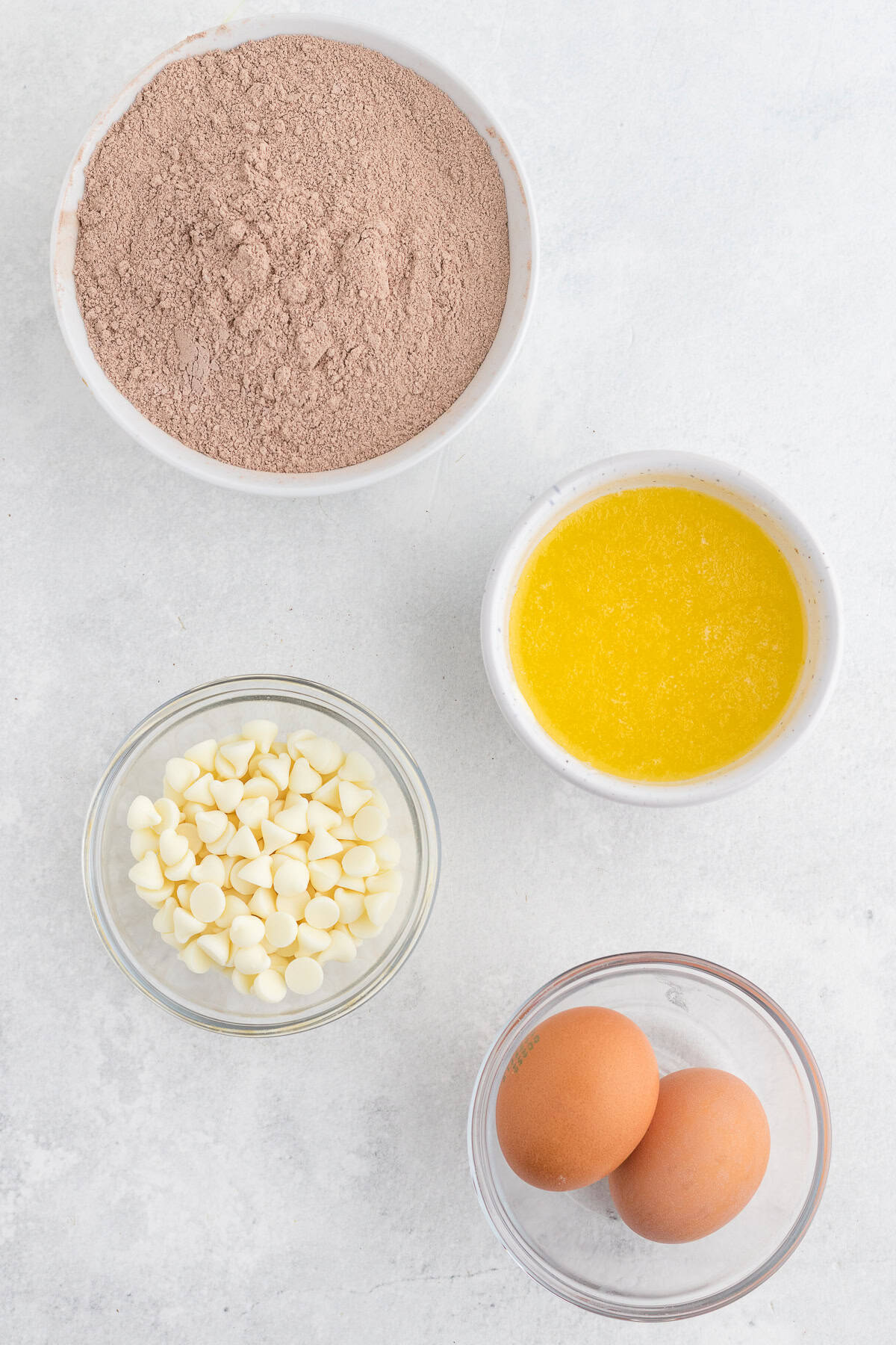 Four ingredients for baking: a bowl of red velvet cake mix, a bowl of melted butter, a bowl of white chocolate chips, and a bowl with two eggs, arranged on a white surface.