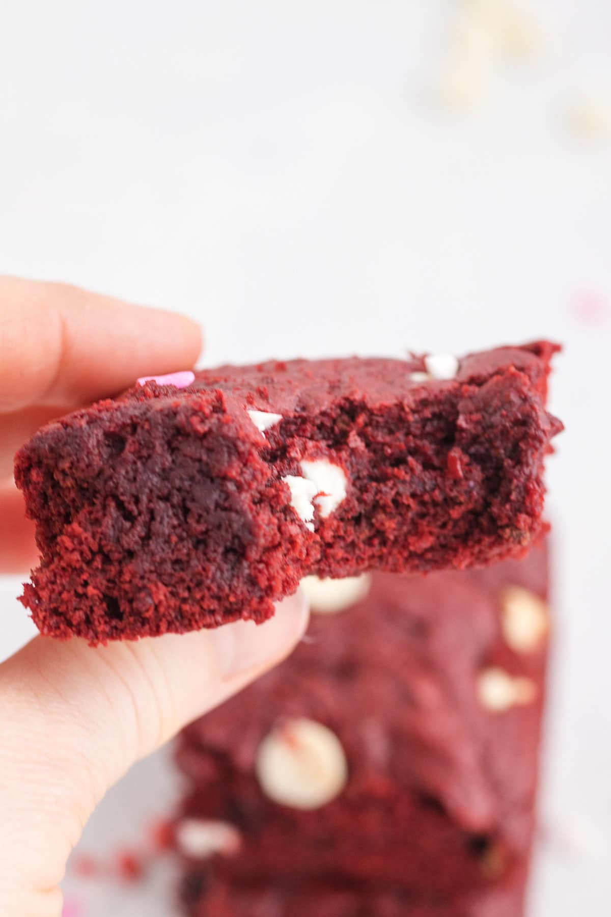 A hand holds a partially eaten Red Velvet Cake Mix Brownie with white chocolate chips, with more brownies blurred in the background.