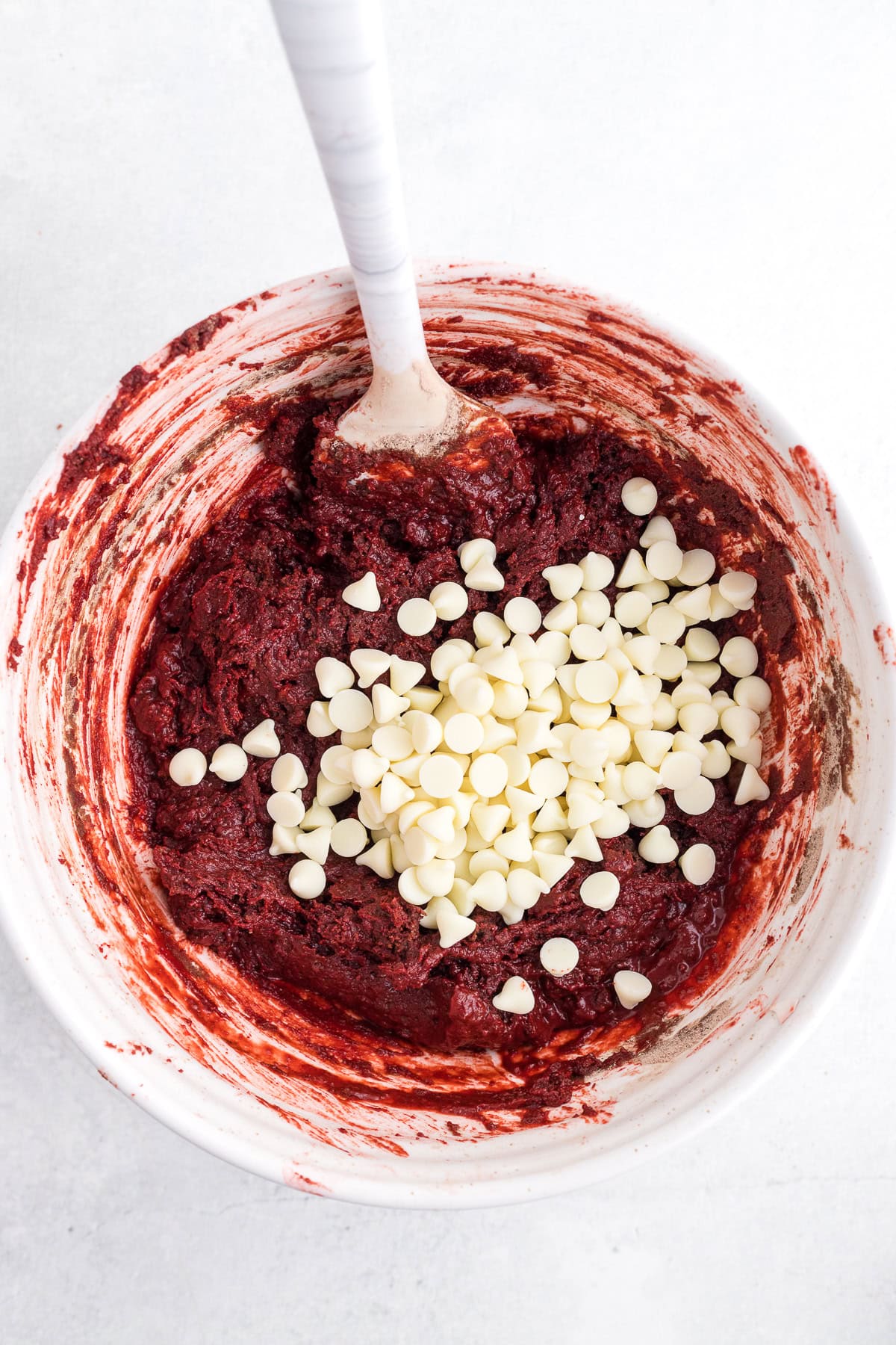A mixing bowl with red dough and white chocolate chips, partially stirred with a white-handled spoon.