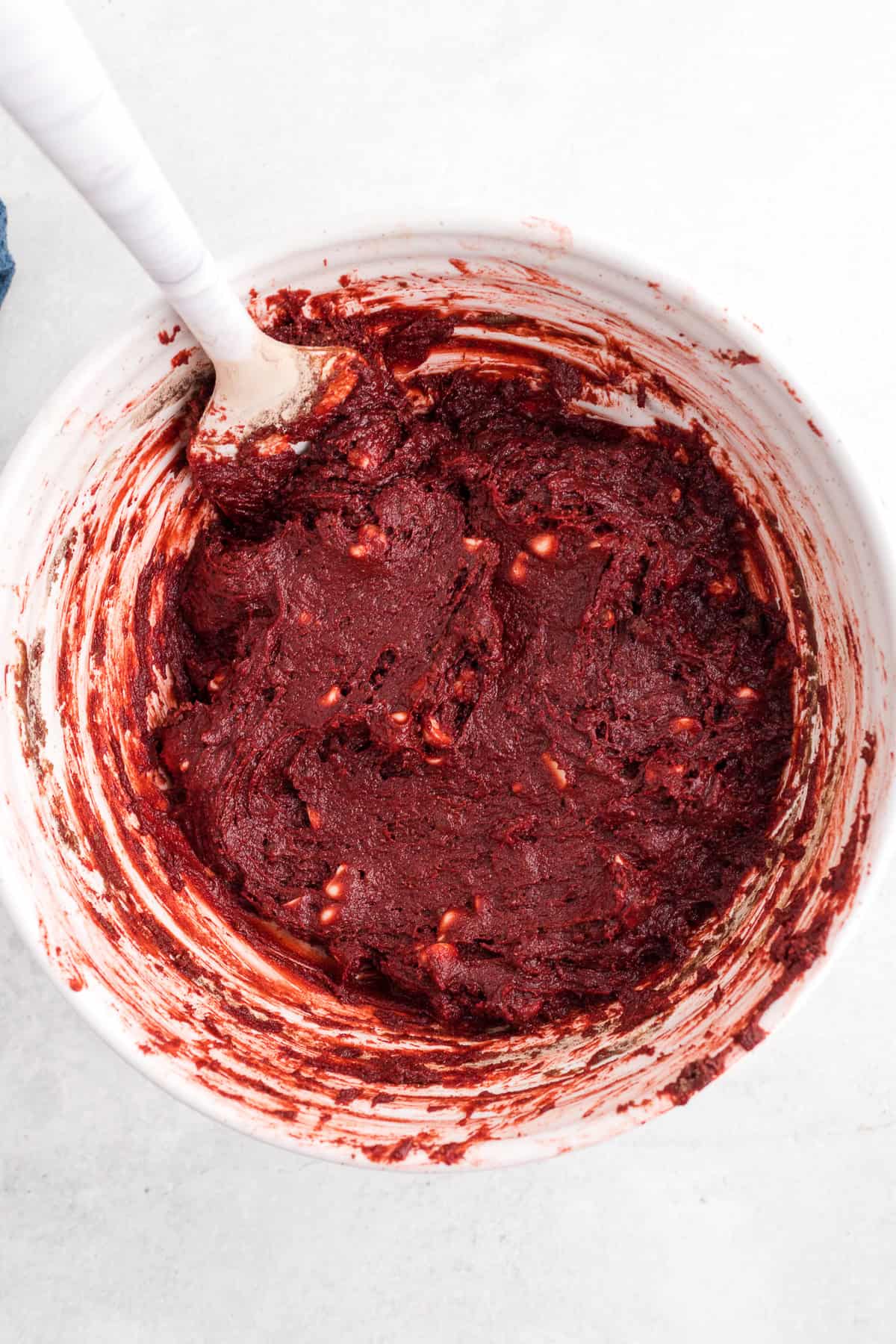 A white mixing bowl filled with thick red dough and a wooden spoon resting inside, viewed from above on a light surface.