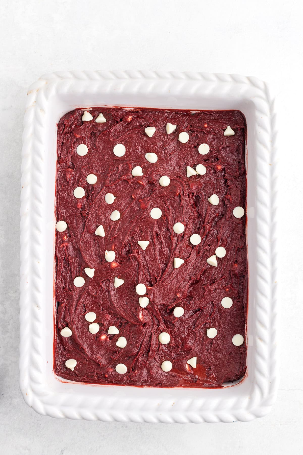 A white rectangular baking dish filled with red batter topped with scattered white chocolate chips on a light surface.