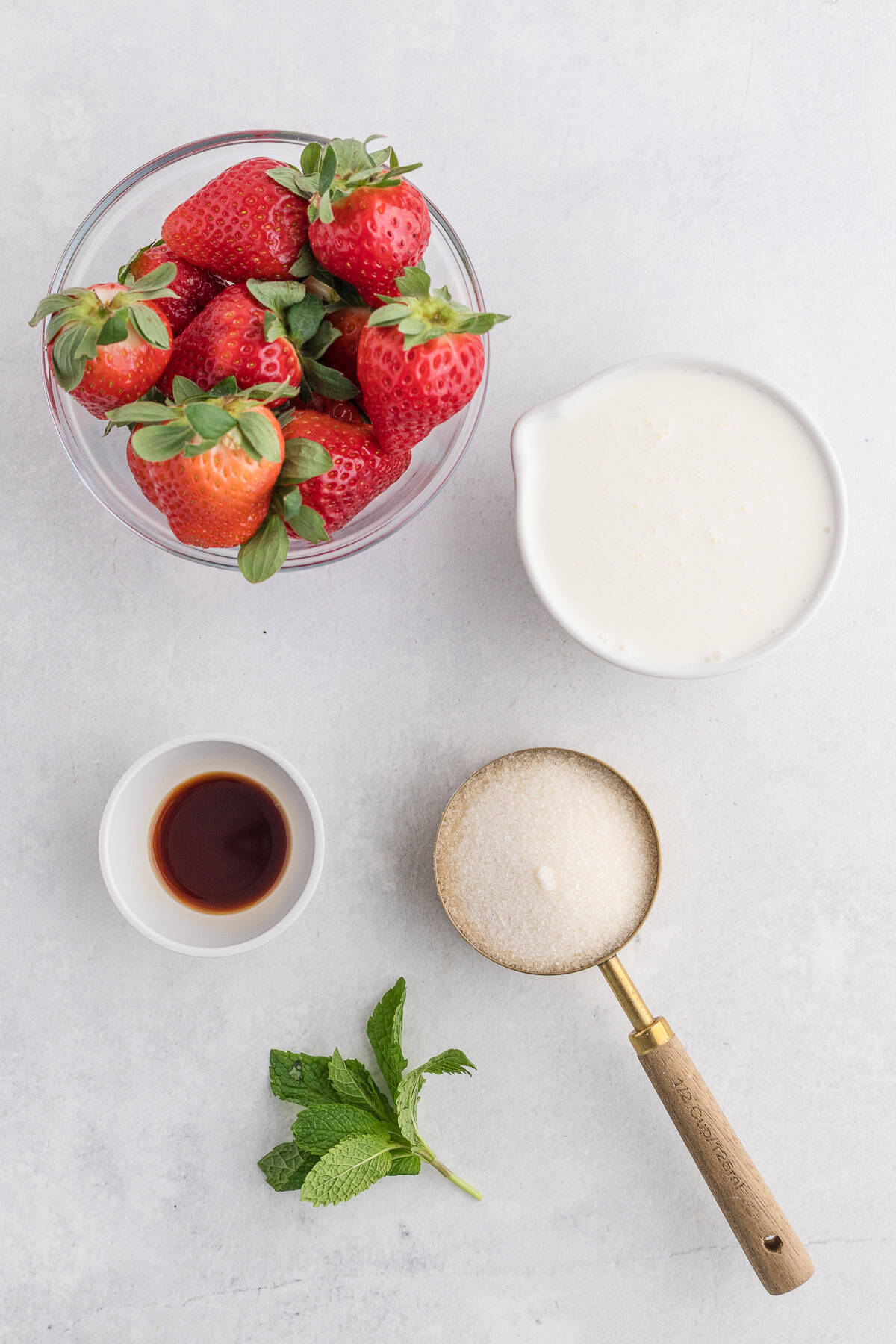 A bowl of strawberries, a cup of cream, a small bowl of vanilla extract, a wooden measuring cup of sugar, and a sprig of mint on a white surface.