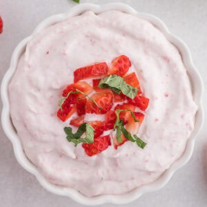 A white bowl filled with pink strawberry mousse, topped with chopped strawberries and fresh green herbs.