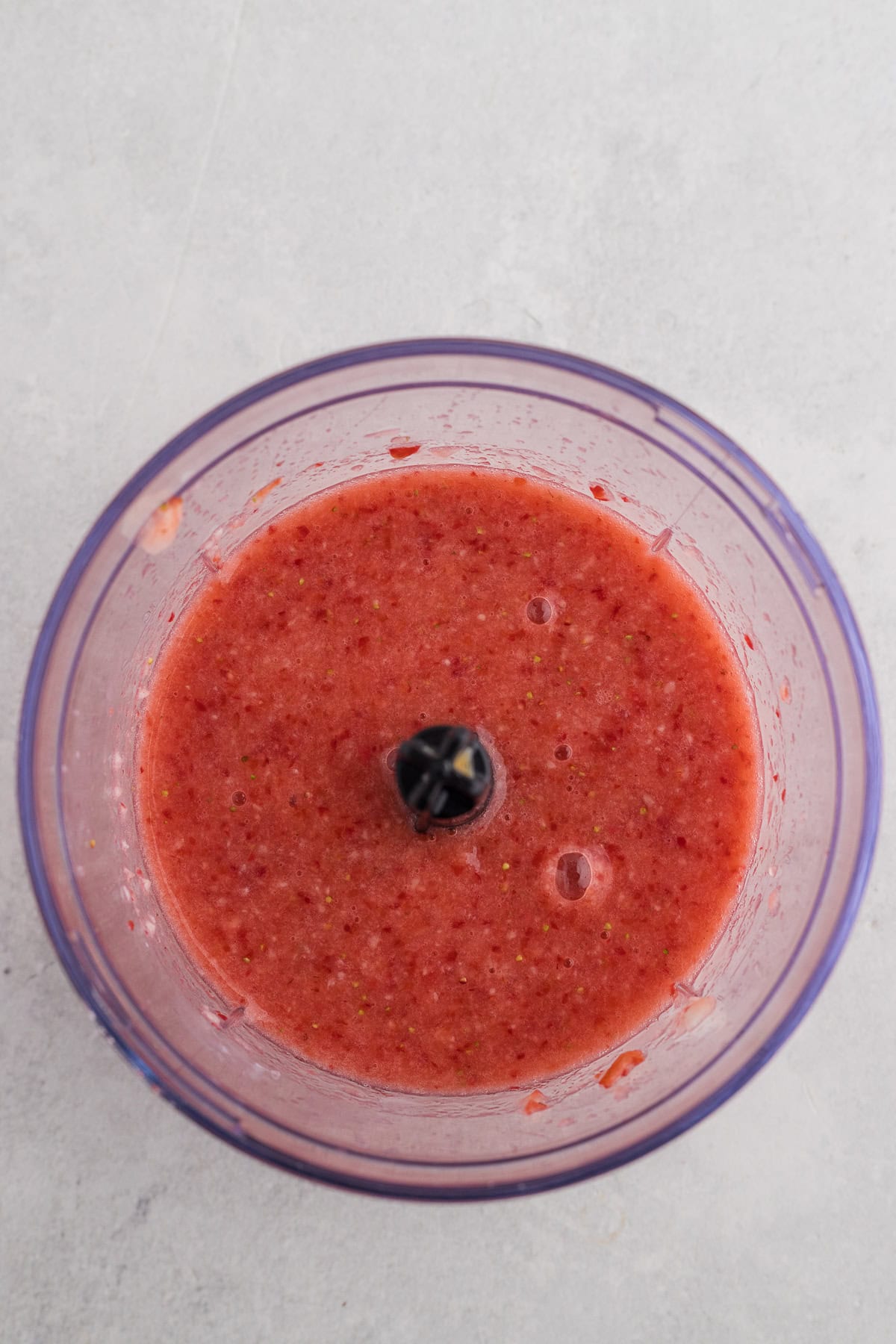A food processor bowl filled with blended strawberry purรฉe, viewed from above on a light gray surface.