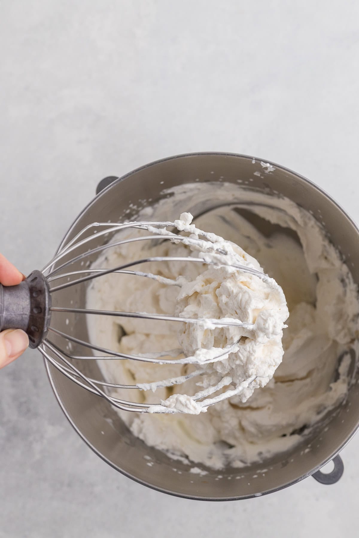 A hand holds a whisk covered in whipped cream above a metal mixing bowl containing more whipped cream.
