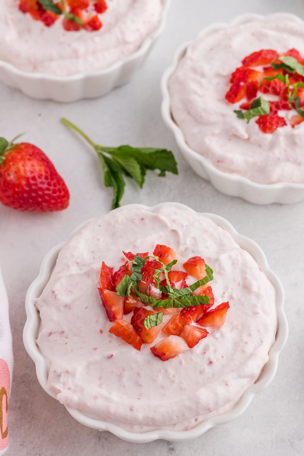 Small white bowls filled with pink strawberry mousse, topped with chopped strawberries and mint, placed on a light surface with a fresh strawberry and mint nearby.
