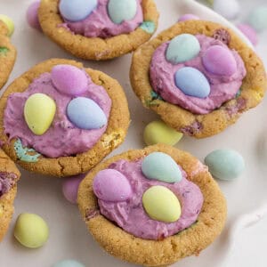 Cookies topped with pink frosting and pastel-colored candy eggs, arranged on a white surface with additional candies scattered around.