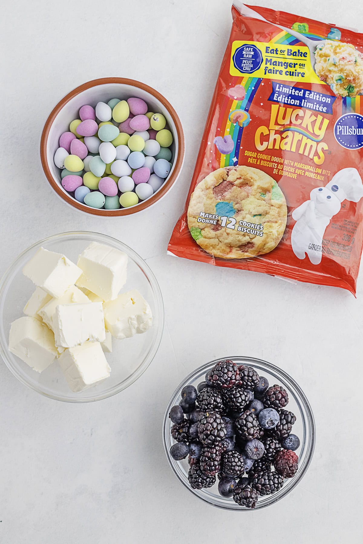 A flat lay of ingredients: a bowl of pastel candy eggs, a packet of Pillsbury Lucky Charms cookie dough, a bowl of cream cheese cubes, and a bowl of mixed blackberries and blueberries.