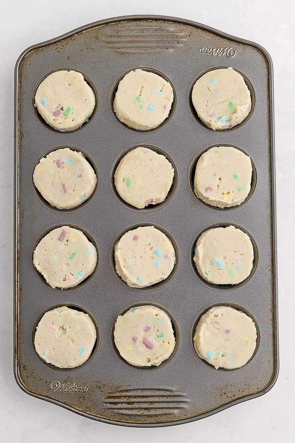 A muffin tin containing twelve portions of raw batter with colorful candy pieces, ready to be baked.