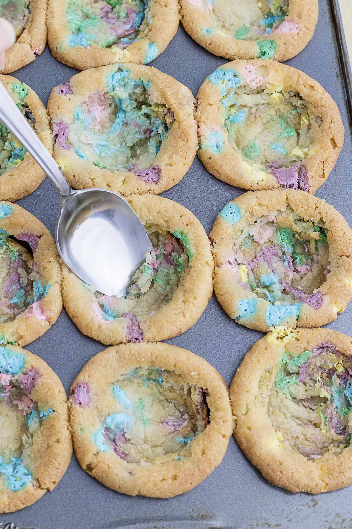 A hand presses a spoon into the center of baked cookies with colorful swirls, creating an indentation in each cookie on a baking tray.
