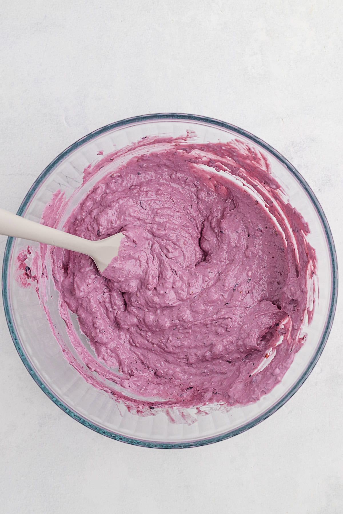 A glass bowl containing thick, bright pink batter being mixed with a white spatula on a light background.