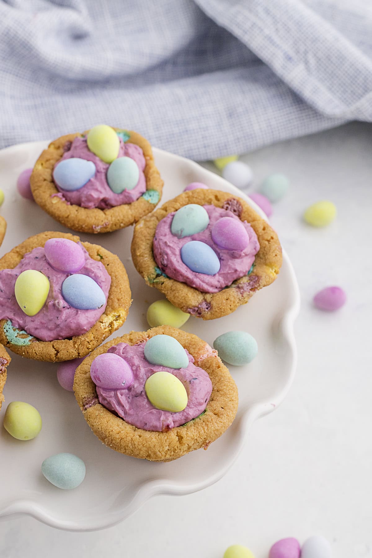 Cookies topped with pink frosting and pastel candy eggs are arranged on a white plate, with additional candy eggs scattered on the white surface nearby.