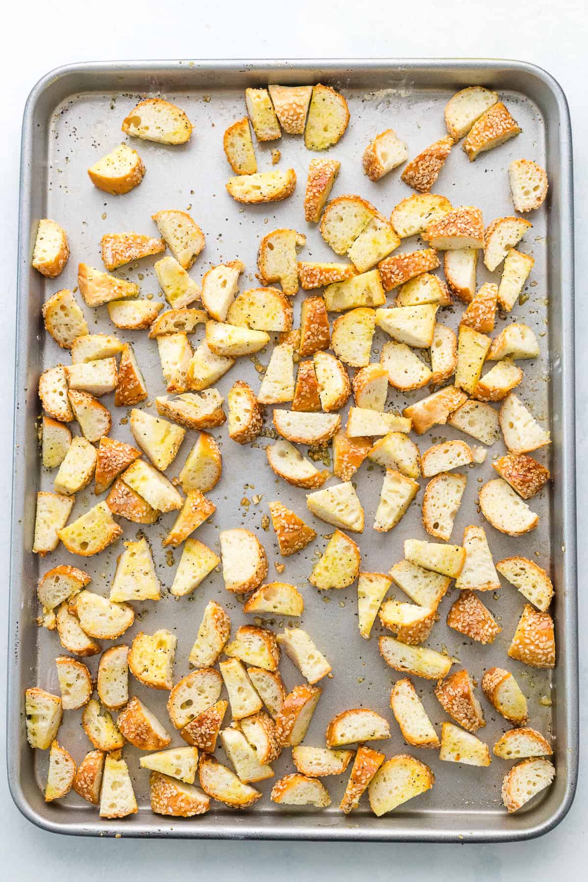 Baking sheet with evenly spread, seasoned bread pieces, toasted and golden brown, on a light background.