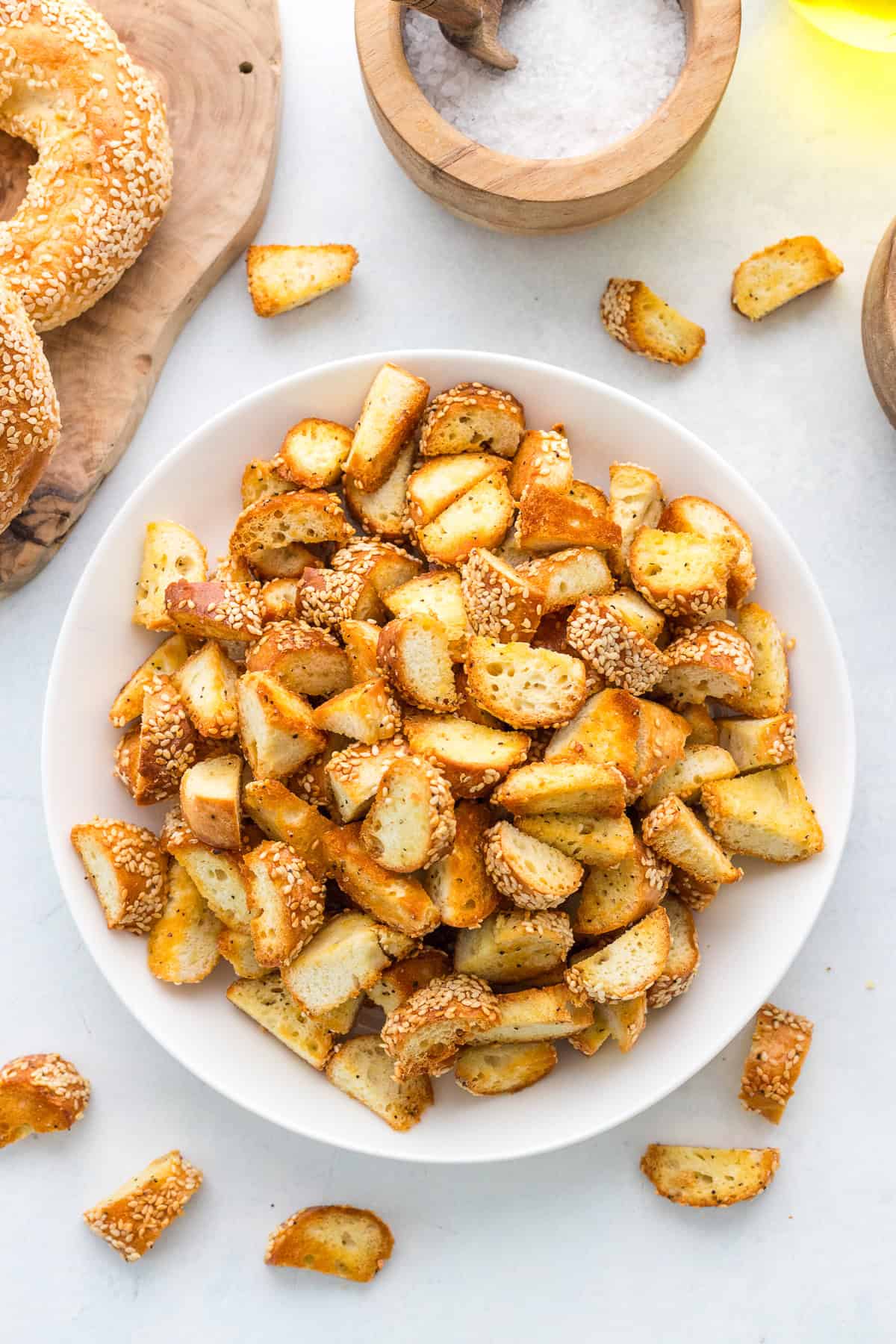 A white bowl filled with sesame pretzel bites sits on a light surface, surrounded by scattered pieces, a wooden salt container, and a pretzel on a cutting board.