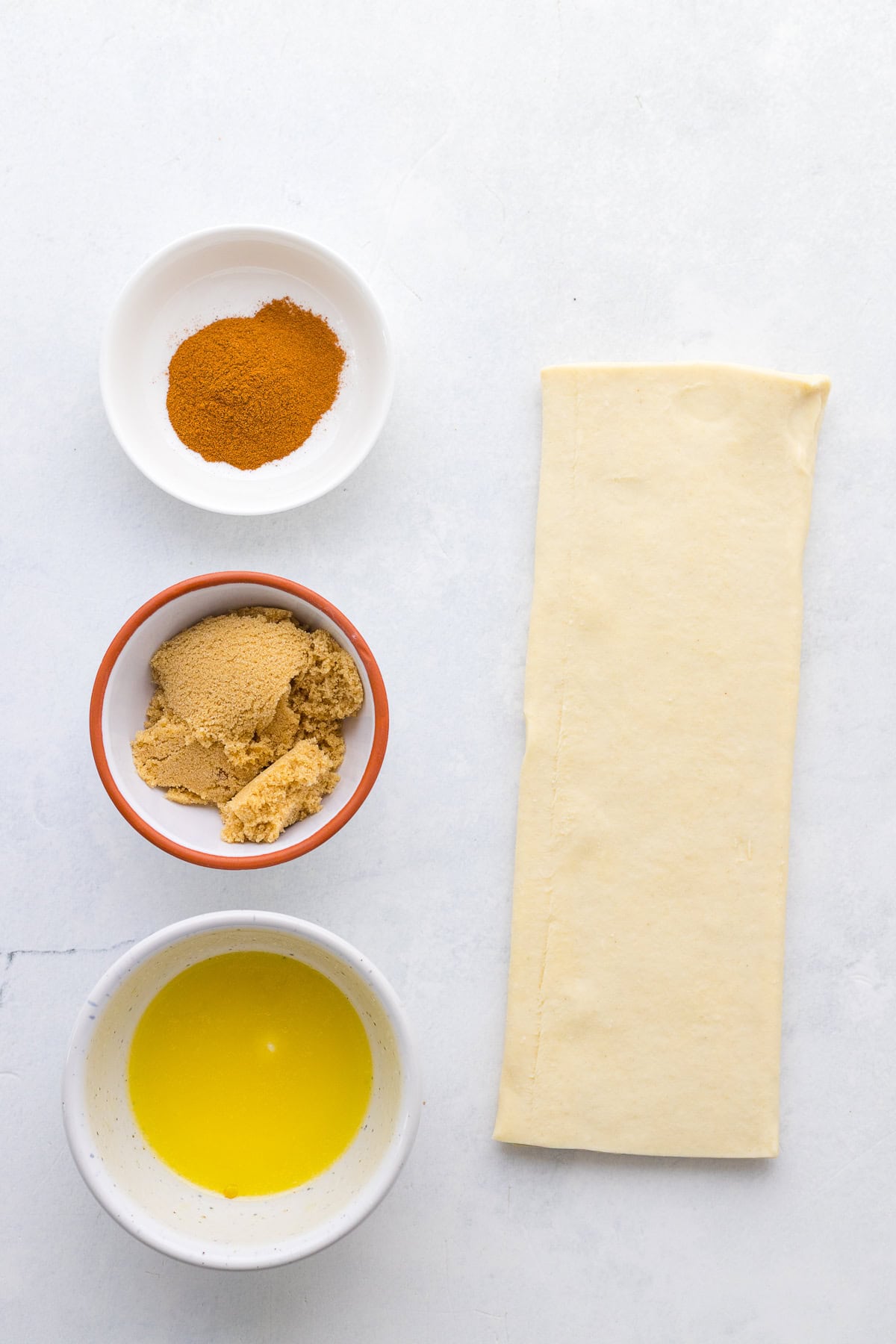 Three small bowls containing ground cinnamon, brown sugar, and melted butter are arranged next to a rectangle of puff pastry dough on a white surface.