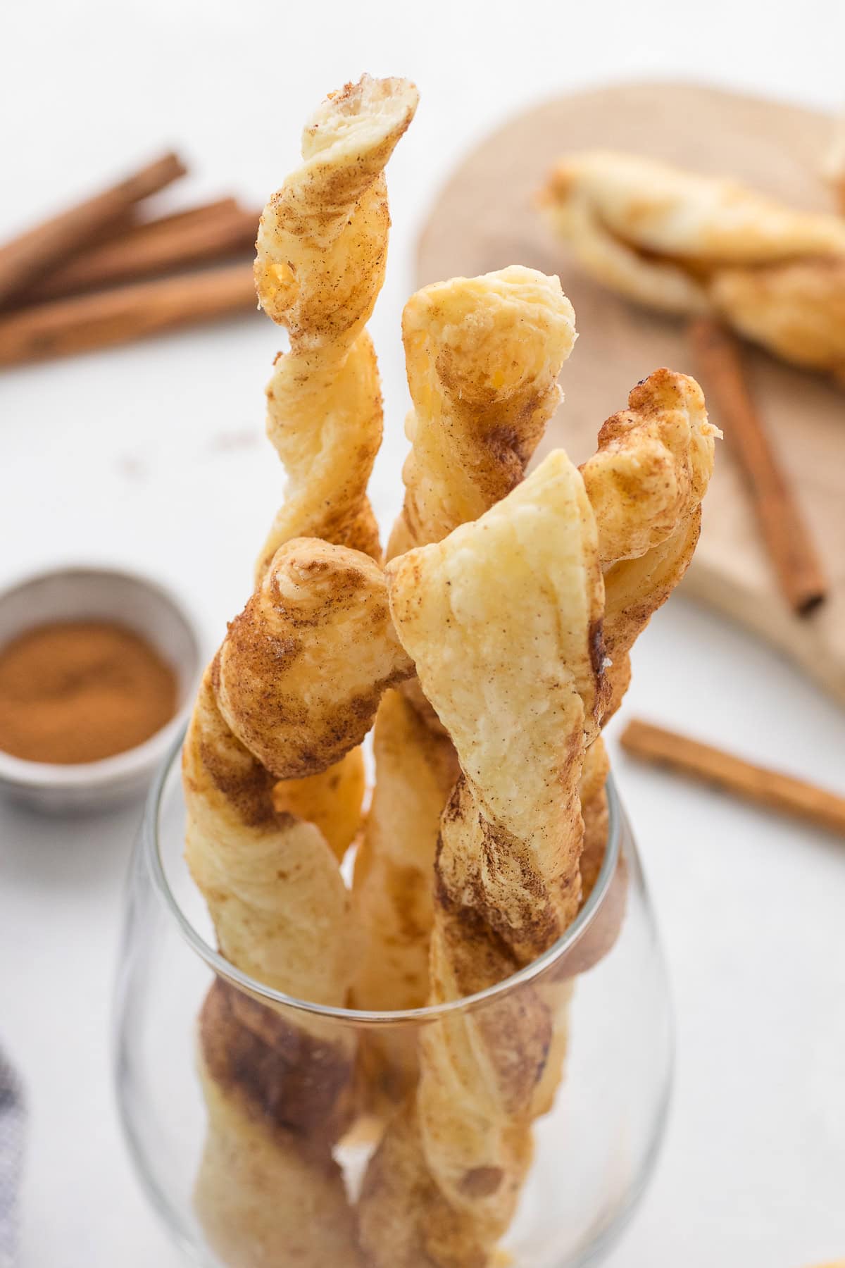 Several twisted puff pastry sticks coated with cinnamon are standing upright in a glass, with more sticks, cinnamon sticks, and a bowl of ground cinnamon in the background.