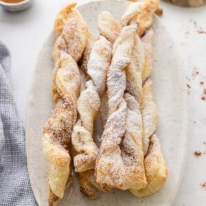 A plate of twisted puff pastry sticks dusted with powdered sugar, arranged on a light-colored oval dish.