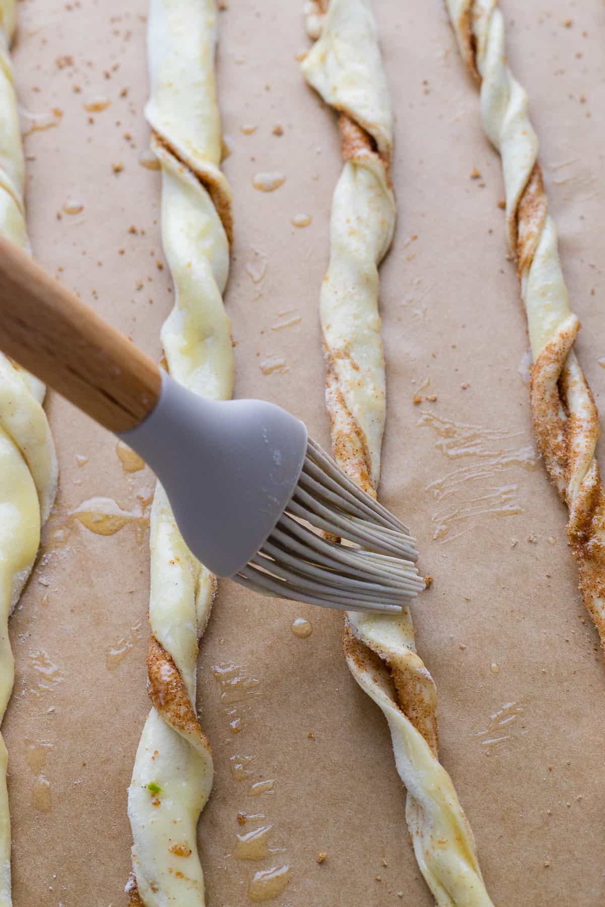 A pastry brush applies egg wash to twisted strips of dough on a parchment-lined baking sheet.