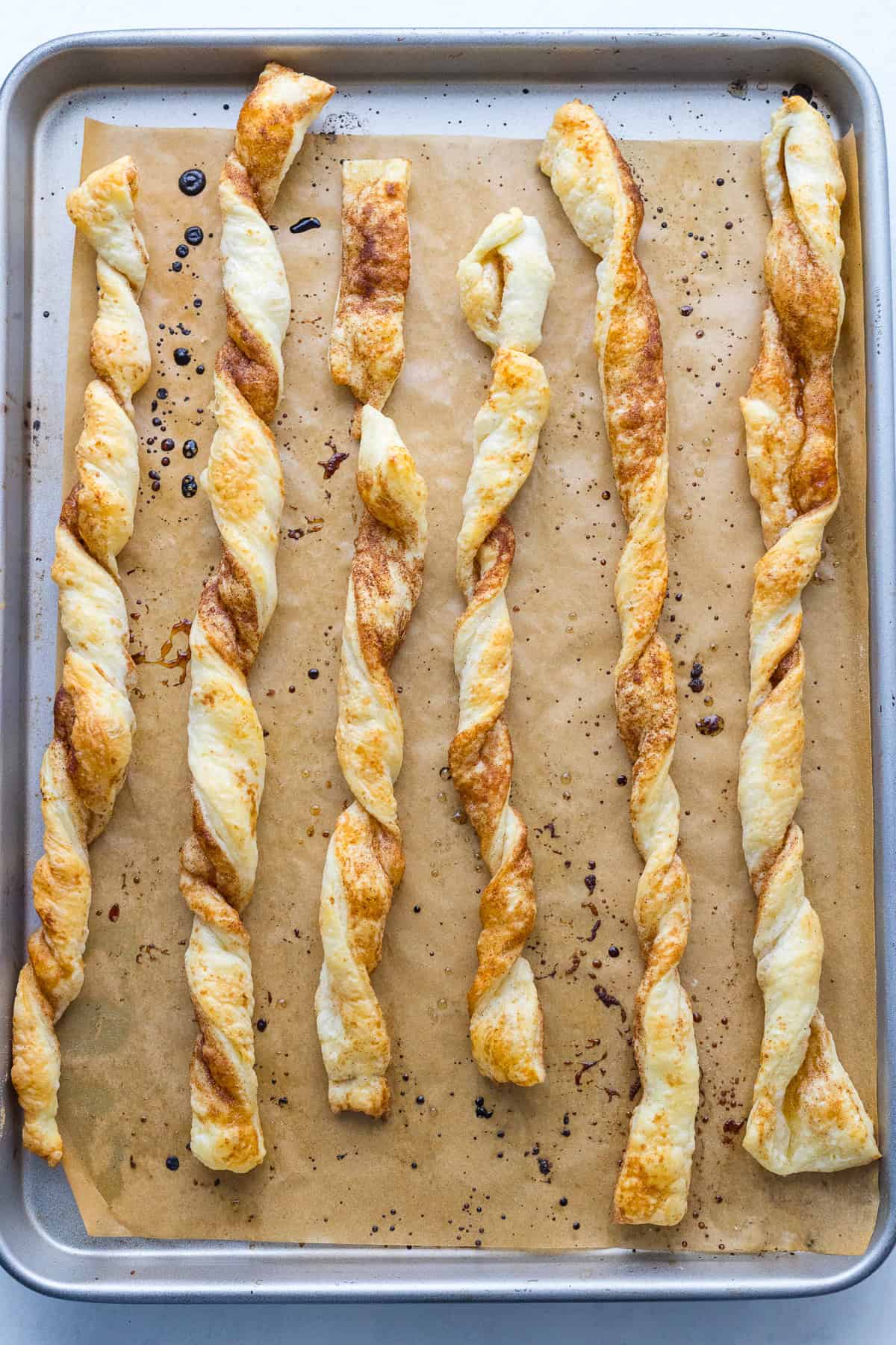 Six baked cheese twists are arranged in a row on a parchment-lined baking sheet.