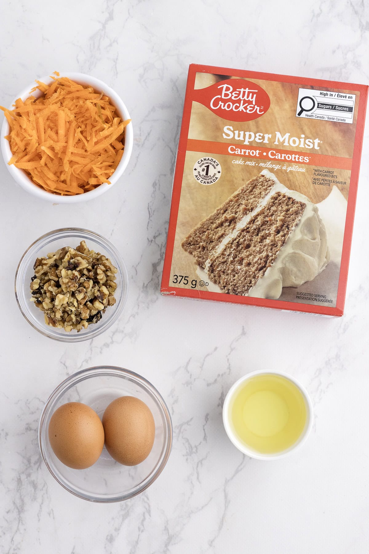 A box of Betty Crocker carrot cake mix, two eggs, a bowl of shredded carrots, chopped walnuts, and a bowl of oil on a marble surface.