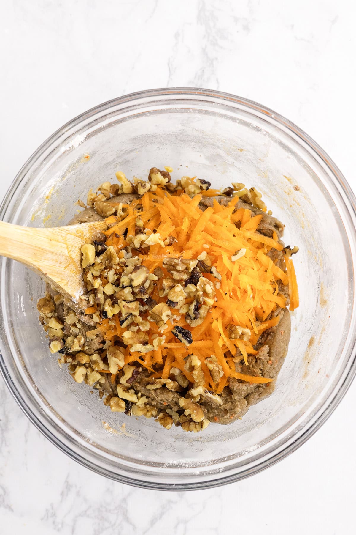 A glass bowl containing a mixture of grated carrots, chopped walnuts, and batter being stirred with a wooden spoon on a white surface.