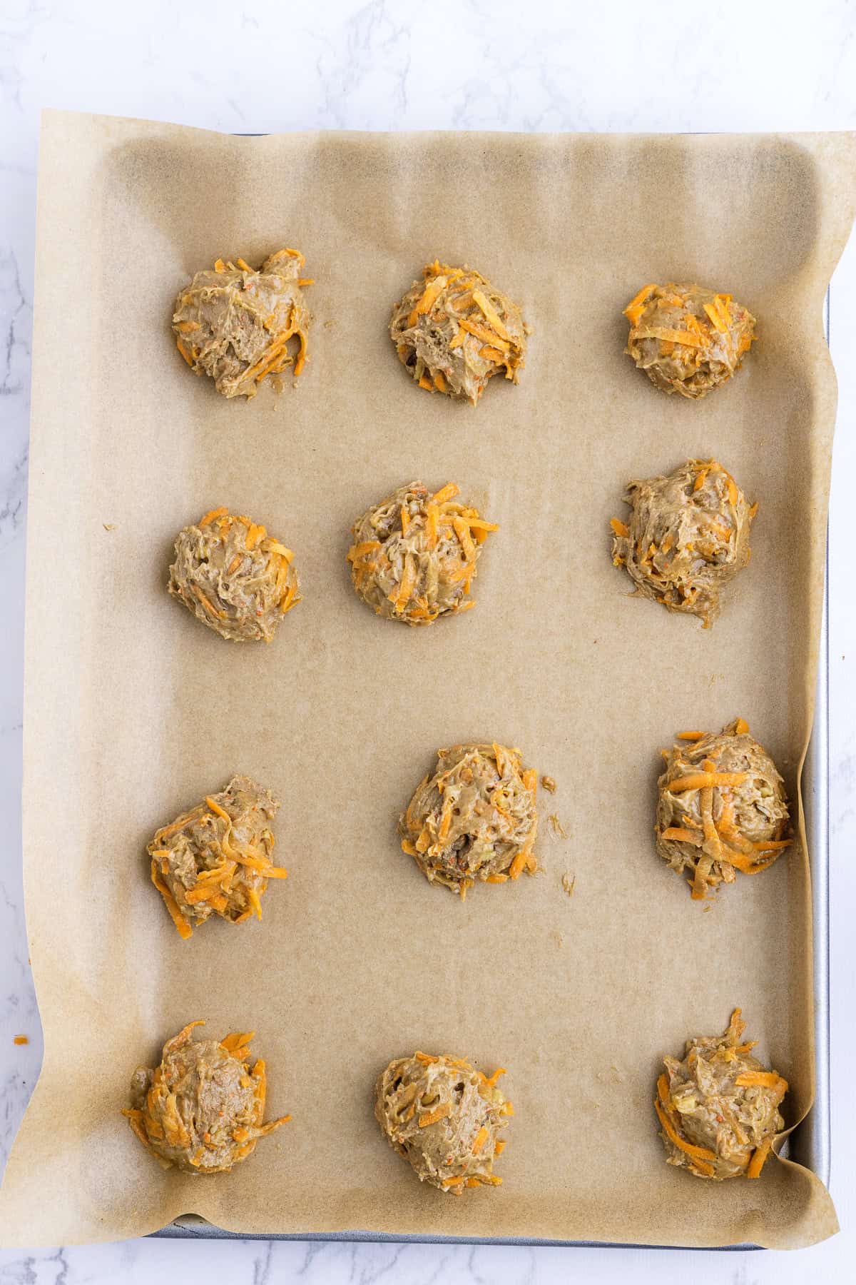 Twelve scoops of raw cookie dough with visible orange shreds are arranged on a parchment-lined baking sheet, ready to be baked.