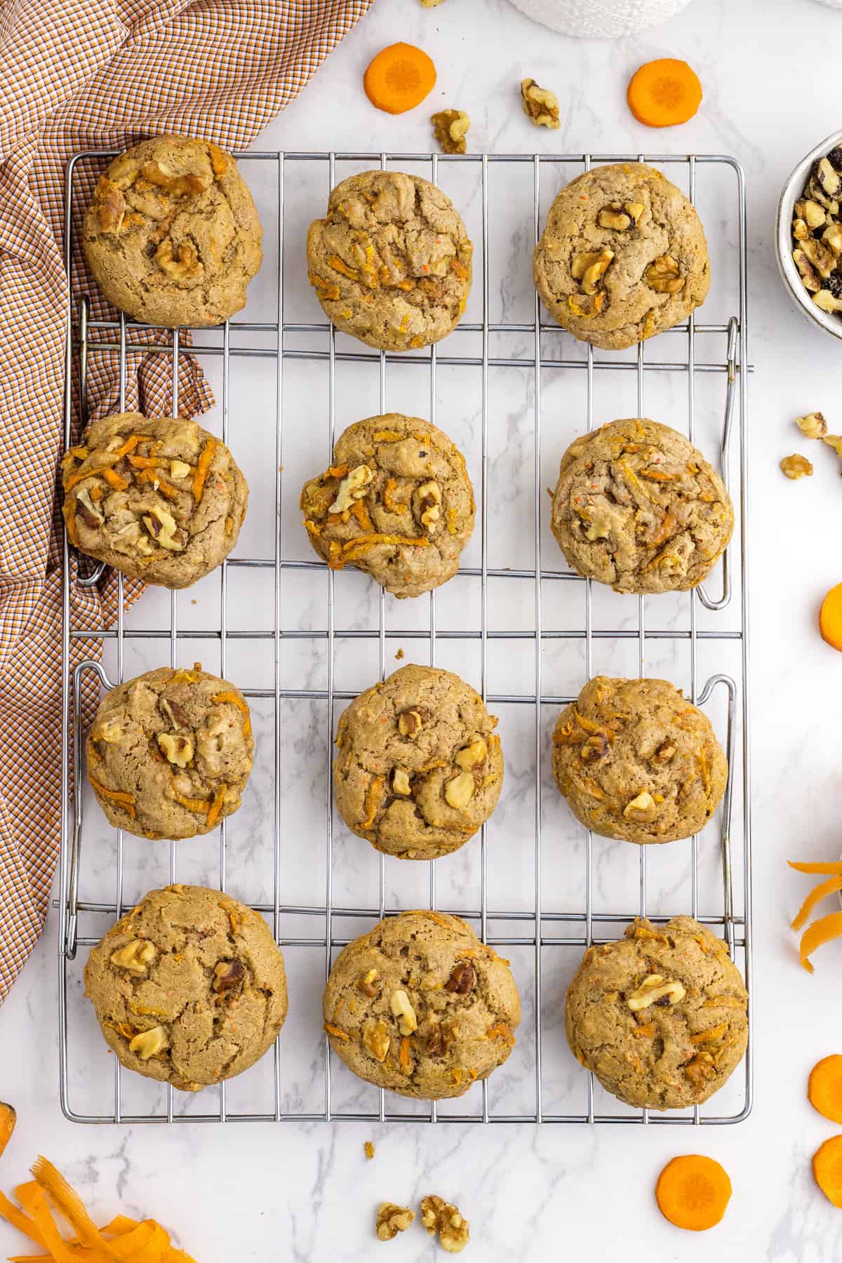 A cooling rack with twelve carrot walnut cookies on a marble surface, with scattered walnut pieces and carrot slices around.