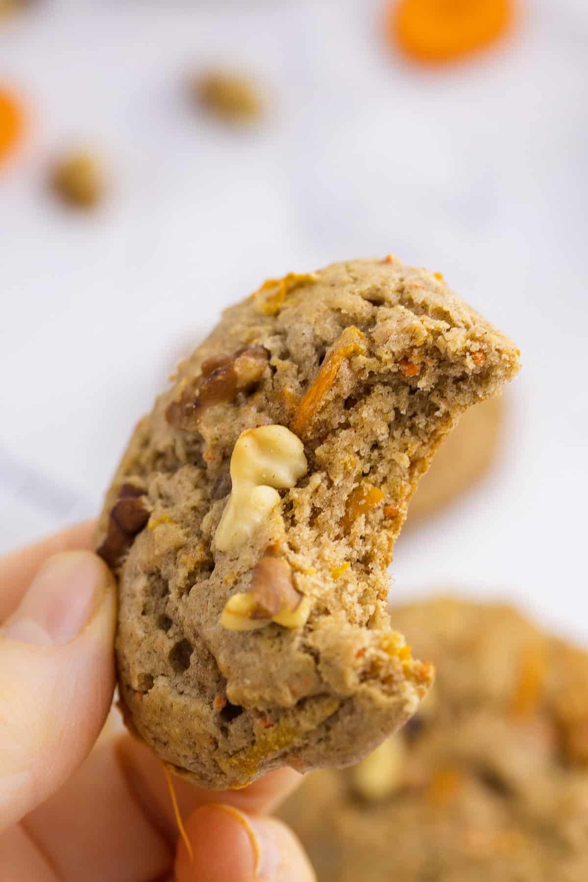 A hand holding a bitten cookie with visible pieces of walnut and shredded carrot.