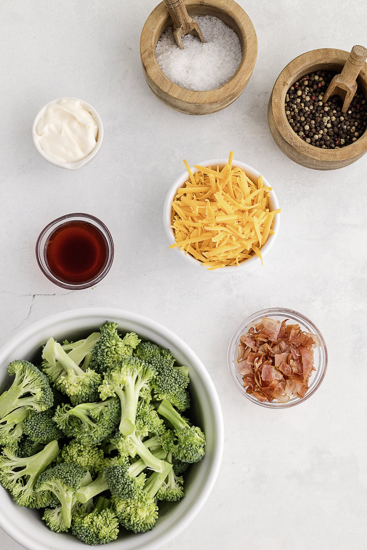 Overhead view of raw broccoli florets, shredded cheddar, crumbled bacon, mayonnaise, red vinegar, salt, and pepper in small bowls on a white surface.