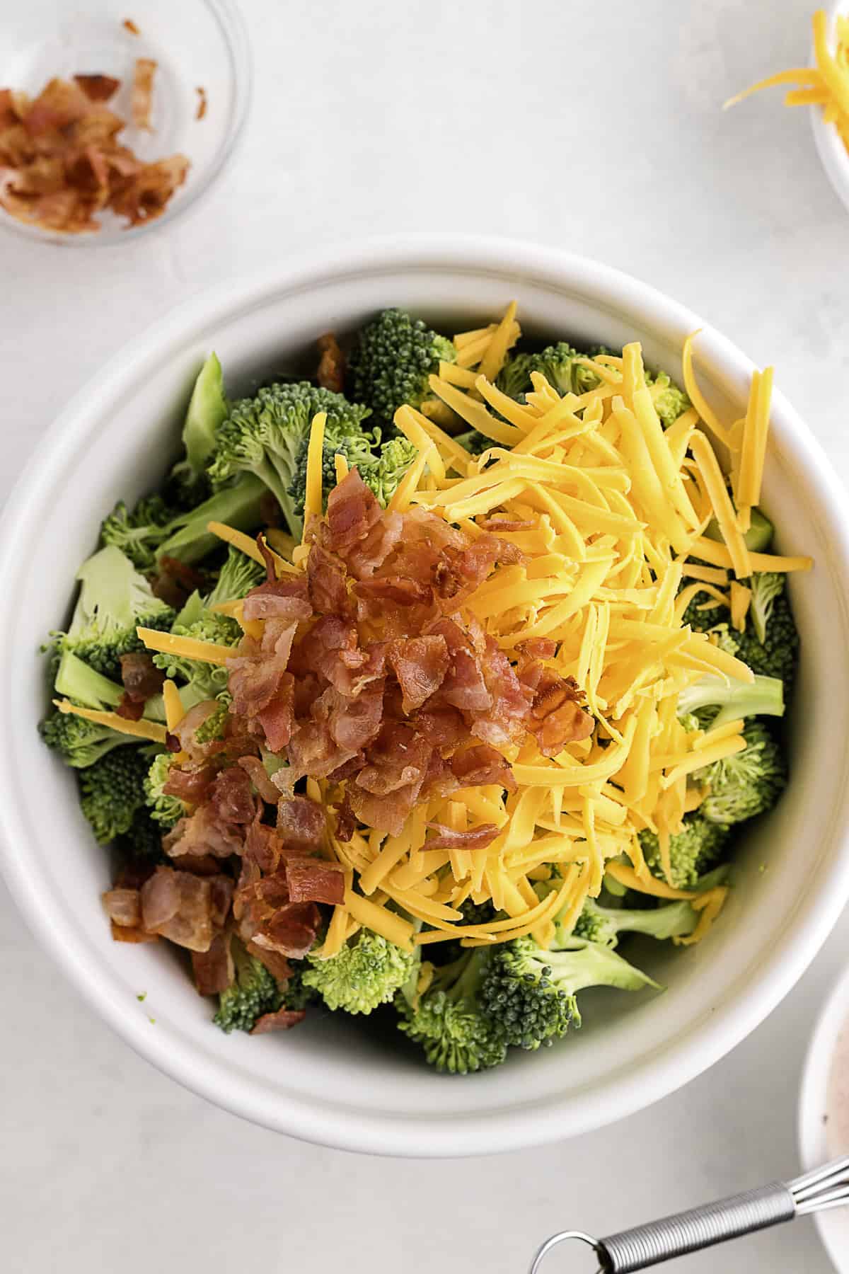 A white bowl filled with raw broccoli florets, shredded cheddar cheese, and chopped cooked bacon.