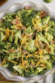Bowl of broccoli salad topped with shredded cheddar cheese and bacon pieces on a light-colored surface.