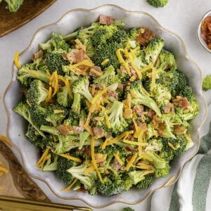 Bowl of broccoli salad topped with shredded cheddar cheese and bacon pieces on a light-colored surface.