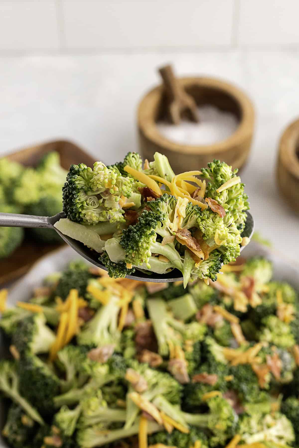 A spoonful of broccoli salad topped with shredded cheddar cheese and bacon bits, held above a bowl filled with the same salad.