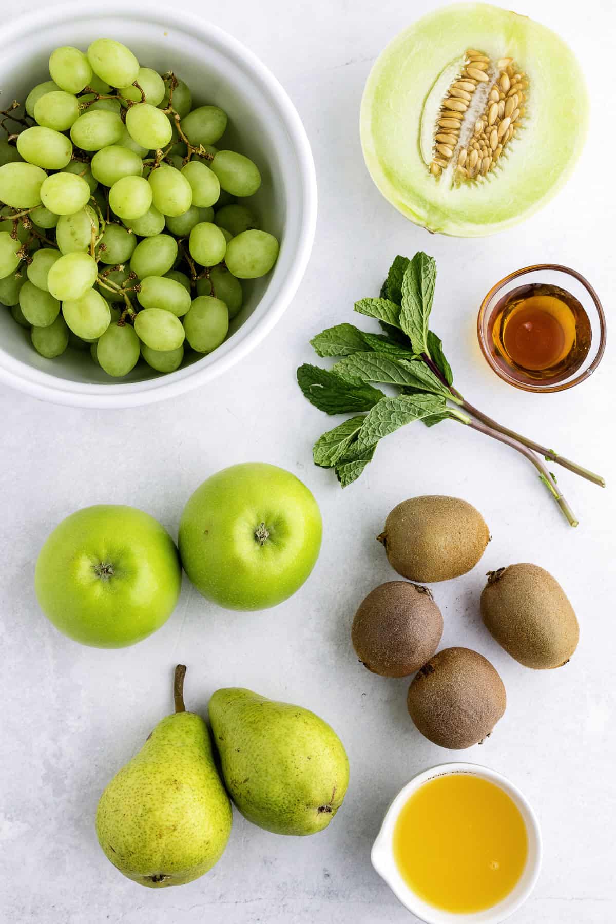 A bowl of green grapes, half a melon, fresh mint, honey, two green apples, three kiwis, two pears, and a small cup of orange juice on a white surface.