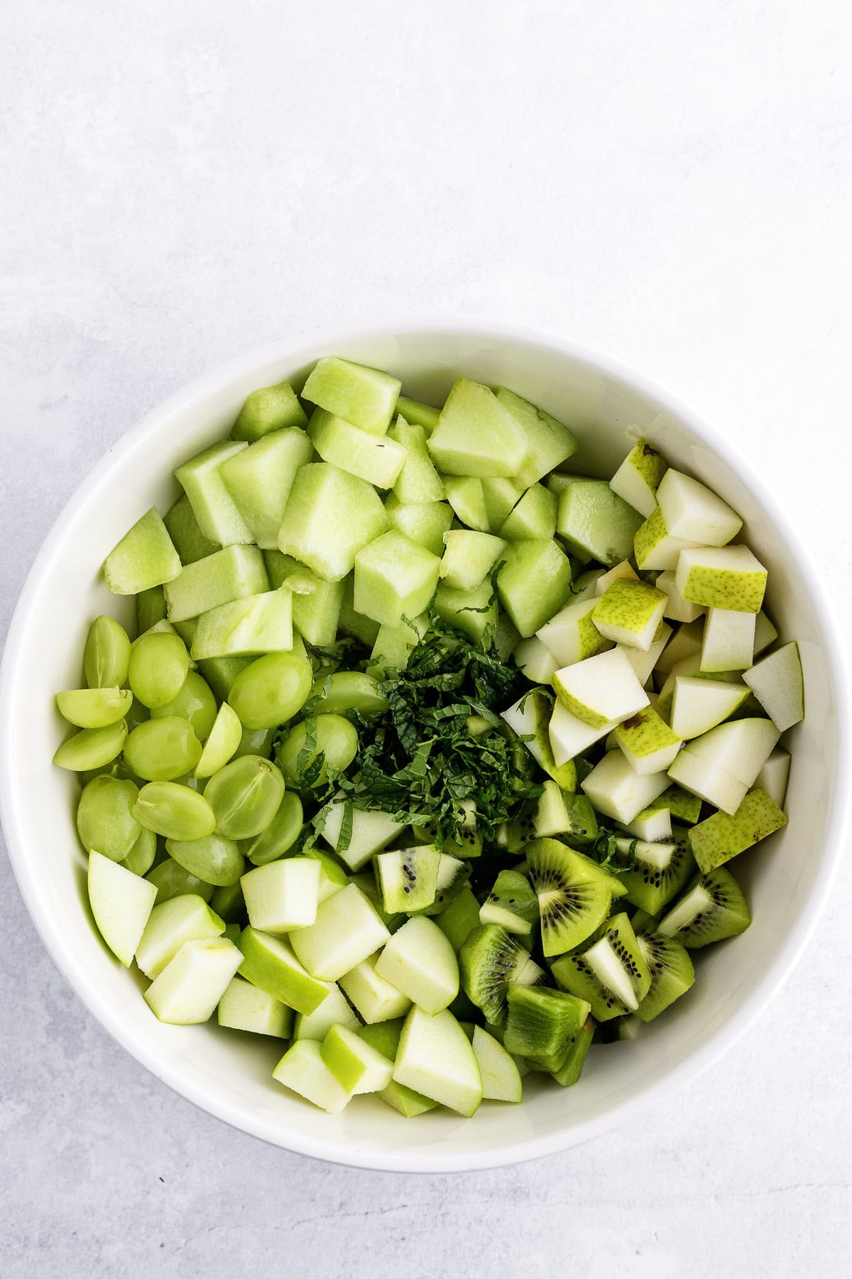 White bowl filled with chopped green fruits including green grapes, apples, pears, kiwi, honeydew melon, and mint leaves, on a light surface.