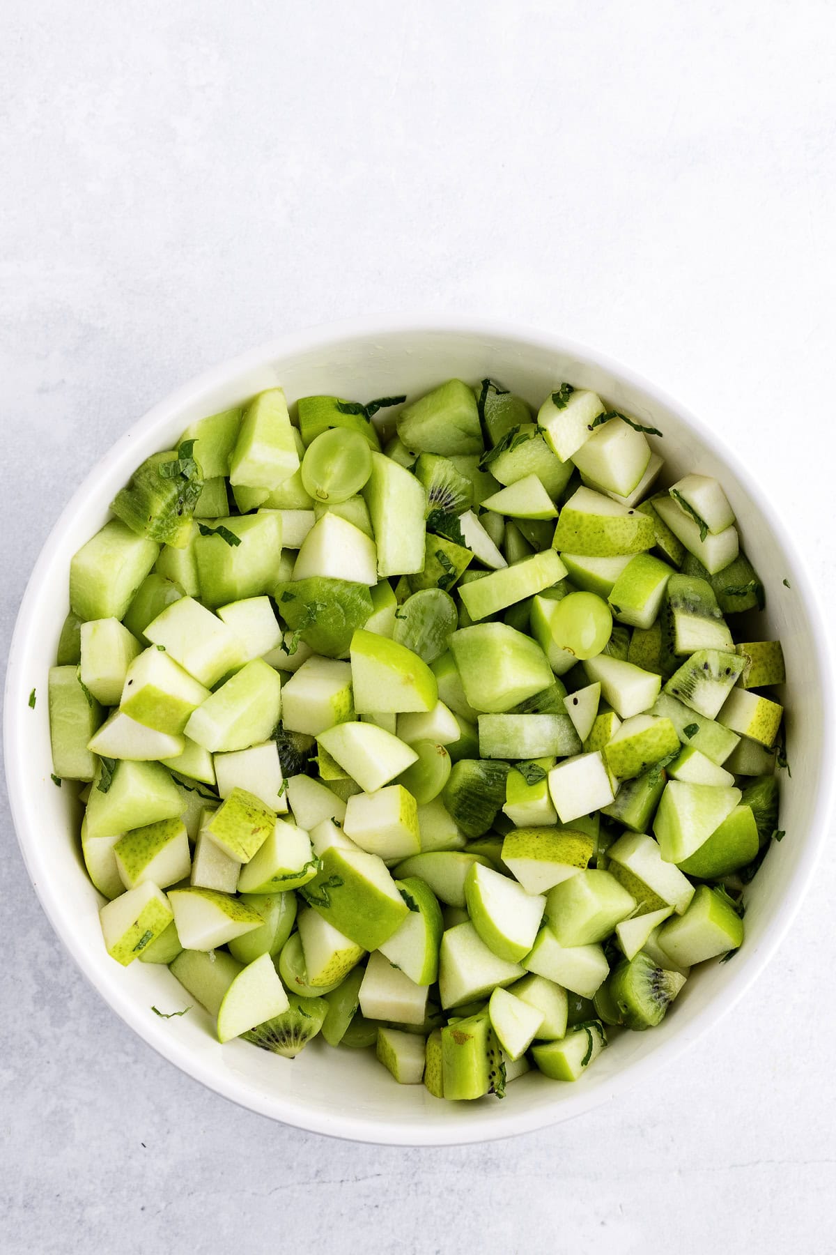 A white bowl filled with chopped green apples grappes, pears and kiwi pieces on a light gray surface.